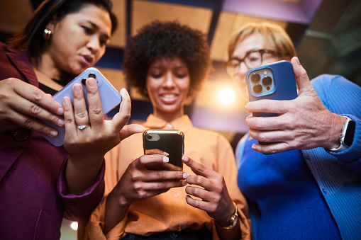 Low angle view of a smiling group of diverse businesswomen checking their smart phones while standing together in an office