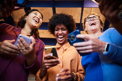 Low angle view of a group of diverse businesswomen laughing at something on their smart phones while standing together in an office