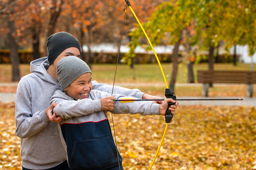 Asian laughing 7 years boy learns to shoot an arrow with the help of his older brother outdoor in park in autumn, close up, selective focus. Family, boys, game, support, having fun, lifestyle concept.