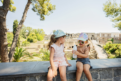 Two baby girl sisters sitting against panoramic view of the ancient city of Pompeii.