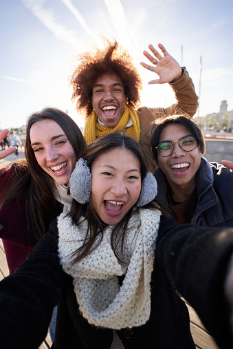 Vertical Selfie of a group of happy people taking selfie photo looking at the camera with a mobile phone in winter clothes. Young smiling mixed race friends having fun in a cold and sunny day.