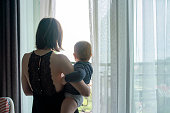 Mother with baby on her hands standing at the window, rear view
