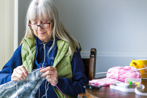 A senior woman in her 70s sits by the window and knits on a rainy day. The window is spattered with raindrops but we can see the fresh green garden outside. The woman is knitting a sweater and is surrounded by knitting apparatus on the table next to her.