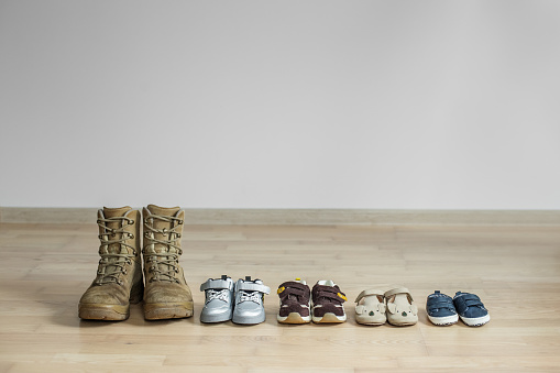 Old worn military boots and lot of baby shoes on wooden floor. Military father and family concept. Copy space