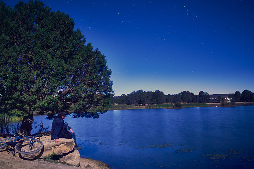 Hombre sentado en soledad en una noche estrellada junto a un lago, arbol en primer plano, soledad