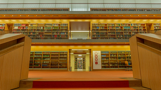 Berlin, Germany-April 2019: The interior design of a massive indoor space of a city library in yellow and orange tones with high ceilings and built-in shelves on the side packed with books and documents. The spectacular architecture of an educational centre in the city.