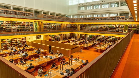 Berlin, Germany-April 2019: The interior design of a massive indoor space of a city library in yellow and orange tones with high ceilings and built-in shelves on the side packed with books and documents. The spectacular architecture of an educational centre in the city.