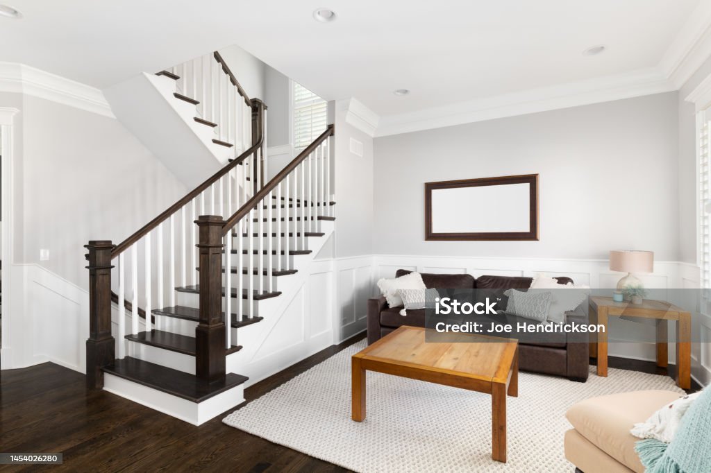 A living room with dark hardwood floors, furniture, and a staircase. A bright, furnished living room near a staircase with wood banisters and steps, white rails, and board and batten walls. Home Interior Stock Photo