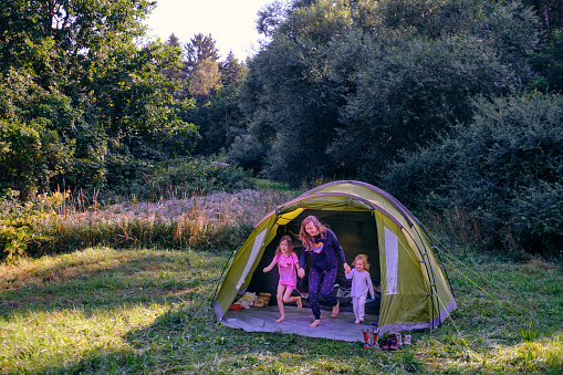 Wild camping with  family, young woman with two small daughters in their pajamas are happily leaving their tent in the morning. Part of happy outdoor moments series.