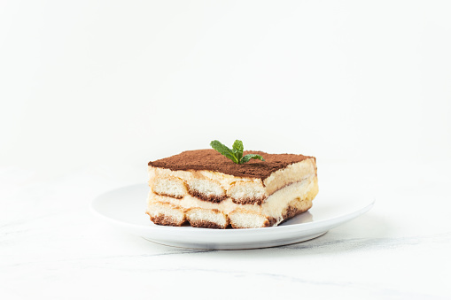Homemade tiramisu cake on white plate against white background. Traditional Italian no-bake dessert made of savoiardi, filled with mascarpone cheese, coffee espresso and sprinkled with cocoa powder
