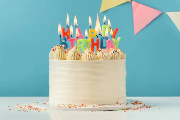 Birthday cake with white cream cheese frosting decorated with multicolored lit happy birthday text shaped candles on a blue background. Tradition of making a wish while blowing out candles on a cake