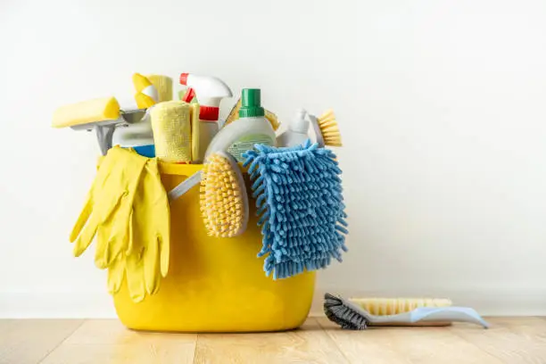 Photo of Brushes, bottles with cleaning liquids, sponges, rag and yellow rubber gloves on white background. Cleaning supplies in the yellow bucket on the wooden floor. Cleaning company service advertisement