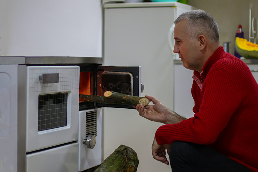 Poor mature man putting firewood in the stove indoors at home, poverty concept.
