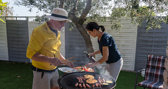 Mature men and woman grilling meat and vegetable in the backyard under the olive tree on sunny day.
