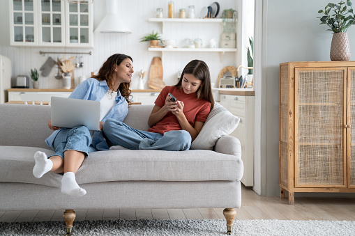 Independent everyday teen girl keeps secrets from mom by hiding phone screen and avoiding surveillance. Smiling young woman with laptop on lap sits on sofa trying to make friends with daughter