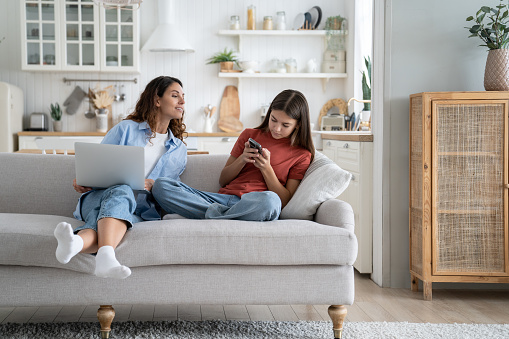 Independent teenage girl trying to hide phone screen from mother sits on sofa. Curious attentive woman looking after daughter who uses internet messengers and making acquaintances in social networks