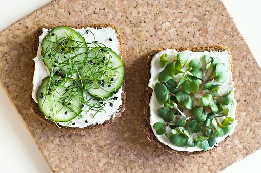 Two type of toasts on whole grain bread with home grown organic microgreens and cucumber on cream cheese. Top view of healthy breakfast on cork board.