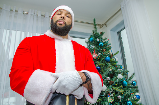 Black Santa, with his real beard posing in front of Christmas tree with serious emotion