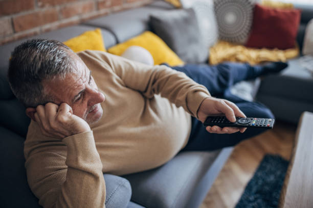 A fat man is lying on a sofa and holding a TV remote