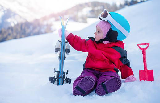 Small girl sit in snow with her first mountain ski in sport outfit including helmet and skiing goggles over mountains