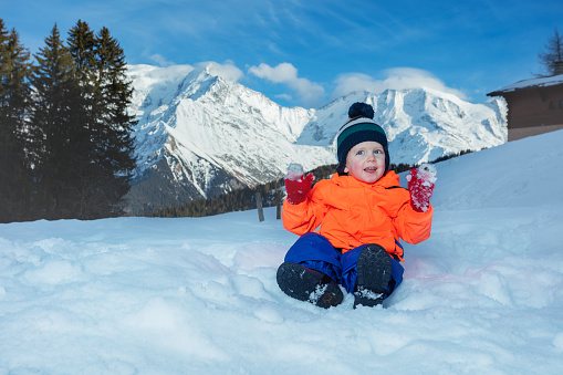 Happy little boy in winter outfit play outside in snow with mountains on background