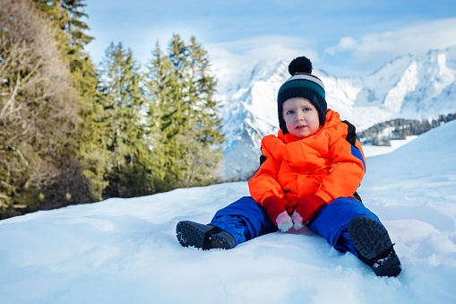 Portrait of a happy young boy sit in winter outfit outside over snow mountains