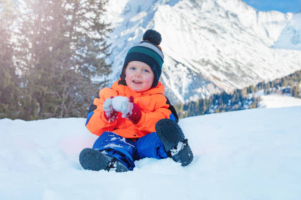 Happy young 2 years old boy sit and making snowball on the snow over mountains