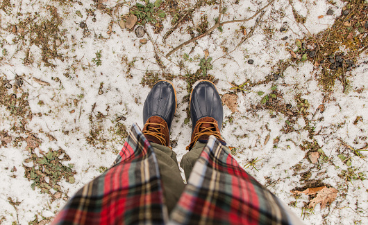 First-Person Downward View of a Woman Wearing Navy & Brown Leather Duck Boots, Green Jogger Pants & a Christmas Tartan Plaid Coat While Standing on Snowy Moss Ground in West Virginia in December 2022.