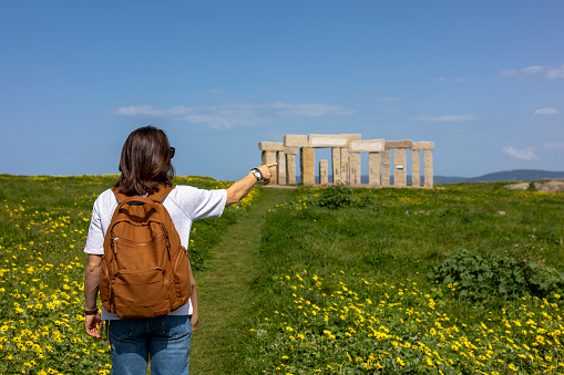 A tourist girl with a brown backpack walks along a green path with yellow flowers to Stonehenge
