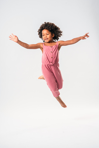 A pretty, multiracial girl in pink onesie, jumping barefoot, high in the air in front of a white studio background. Arms stretched laterally, one leg bent up in the knee, looking away.