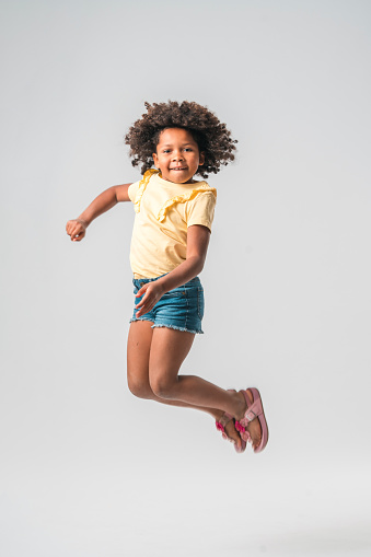 Little black-American girl in summer clothes, jumping high up in the air with bent legs, arms helping lift the body. Wearing pink sandals. Looking at the camera with a toothy smile and her hair flying up.