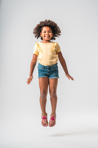 Little African girl in summer clothes, jumping high up in the air with extended legs, wearing pink sandals. Looking at the camera with a toothy smile and her hair flying up.