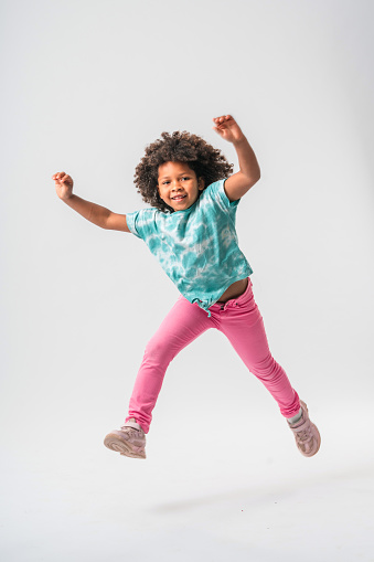 Pretty, school-aged multiracial girl in the middle of a long running leap. Both feet are off the ground, arms up in the air, away from the body. Looking confidently at the camera. Wearing pink trousers and sport shoes.
