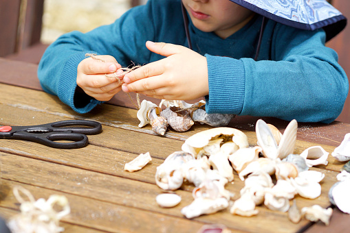 Young boy makes Christmas decorations with seashells he has collected from the beach. He threads them onto a piece of string then ties a knot to make the decorations to hang on the tree.