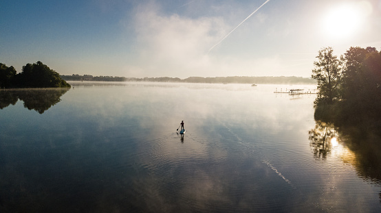Woman paddling on SUP board on beautiful lake aerial drone view with reflections from above. Standing up paddle boarding adventure in early morning sunrise. Germany lake district Mecklenburg.