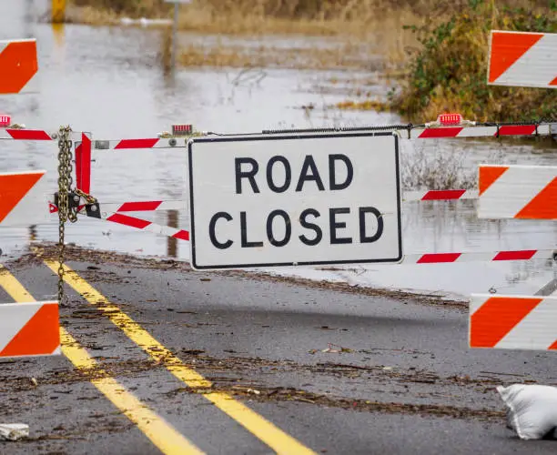 A flooded road with a Road Closed sign and barricade. The barricade is chained with a lock so drivers will not move it to drive around.