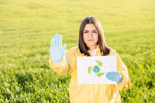 Person in overalls holds paper with a call to save the planet while standing on green field on sunset and the other hand shows a gesture to stop and not pollute the planet. Concept of bad ecology and environmental protection