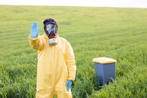 A woman in a yellow protective suit stands in the middle of a green field in a protective gas mask and shows a gesture to call to stop and save the planet. A woman stands near the garbage can and calls to save the planet