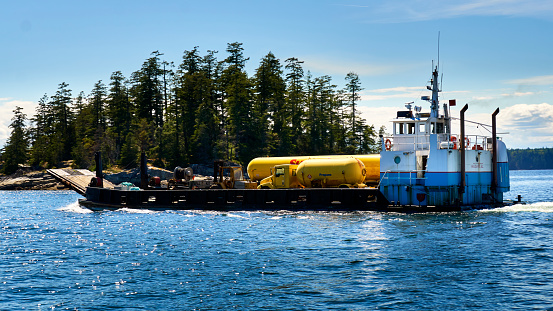 Campbell River, Canada – July 05, 2020: A closeup of a self propelled barge delivering propane, equipment and a yellow propane truck moving forward on choppy ocean waters.