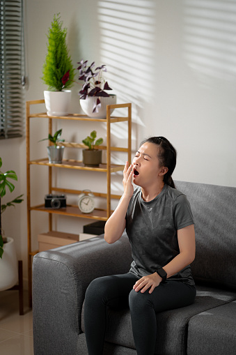 Asian woman feel tired exhausted from exercised sitting and yawning on sofa in living room at home.