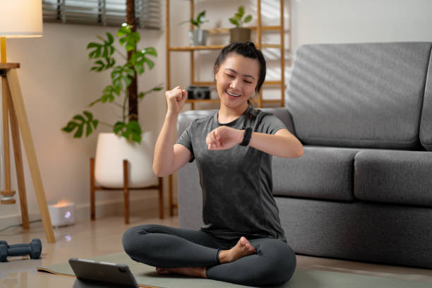 スマートウォッチを見て、トレーニングの目標が成功したことに満足しているアジアの女性は、自宅のリビングルームでリラックスした時間。 - living room learning healthy lifestyle one person ストックフォトと画像