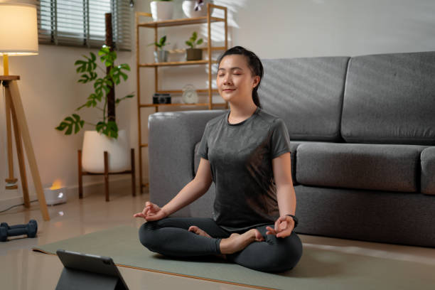 アジアの女性は、自宅のリビングルームでデジタルタブレットを使ってオンラインでヨガのクラスを学ぶヨガの練習の祈りの姿勢でマットの上に足を組んで座る。 - living room learning healthy lifestyle one person ストックフォトと画像