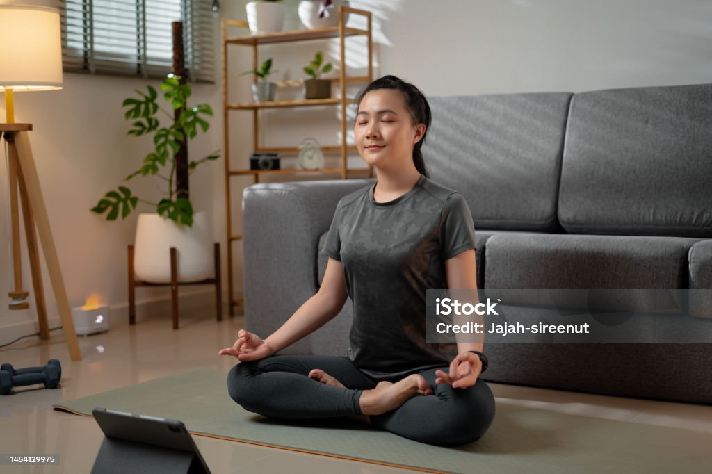 Asian woman sit cross-legged on mat in prayer position of yoga practice learning yoga class online with digital tablet in living room at home. Asian woman sit cross-legged on mat in prayer position of yoga practice closed eyes breath deep feel comfort do meditation practice learning yoga class online with digital tablet in living room at home. Relaxation Stock Photo