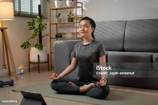 Asian Woman Sit Crosslegged On Mat In Prayer Position Of Yoga Practice Learning Yoga Class Online With Digital Tablet In Living Room At Home Stock Photo - Download Image Now
