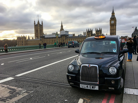 London, UK - Nov 2016: London taxi stopped by pedestrian walkway on Westminster Bridge waiting for passenger with Westminster Palace and Big Ben clock tower and cloudy sky in background.