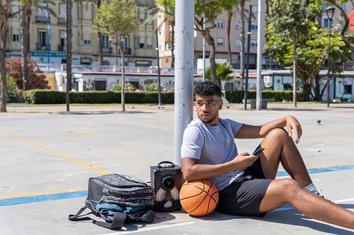 Portrait of a young muslim basketball player resting holding a smartphone on a city court, urban spo