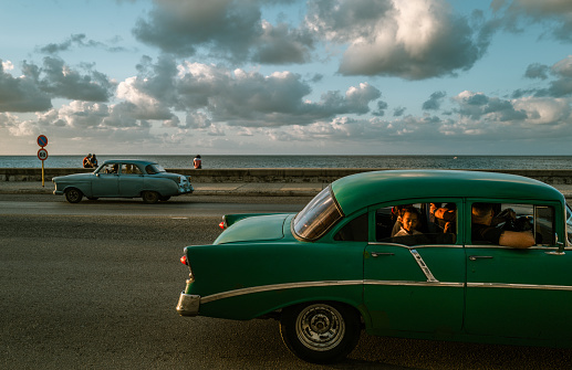 Havana, Cuba - December 9th 2022: Classic vintage cars driving along the Malecon at old Havana during dusk hour. Sunset lighting the face of a cuban girl in the car. Malecon is iconic spot for tourists, also the most popular place for cubans to hang out.