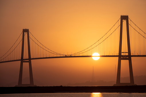 A high angle shot of the bright sun setting behind a big bridge over the sea in South Korea