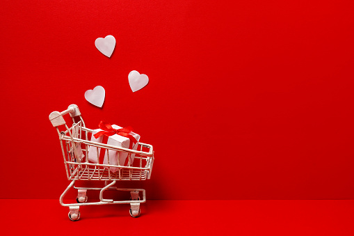 Shopping trolley with gift box, white love hearts on red background. St. Valentine's Day shopping and sale. copy space