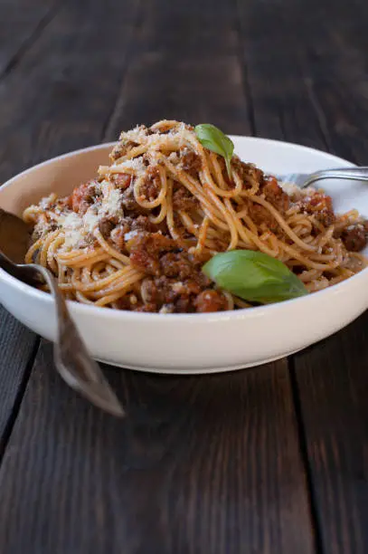 Traditional homemade spaghetti bolognese with grated parmesan cheese and basil leaves. Served in a white bowl or plate with rustic fork and spoon isolated on dark wooden table background with copy space